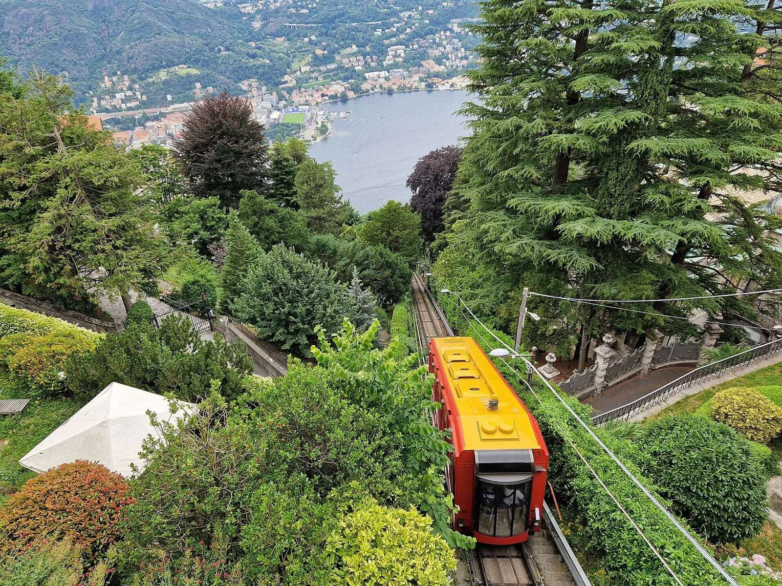 Vettura rossa e gialla della funicolare Como-Brunate sul binario tra il verde, con il primo bacino del lago e la città di Como sullo sfondo