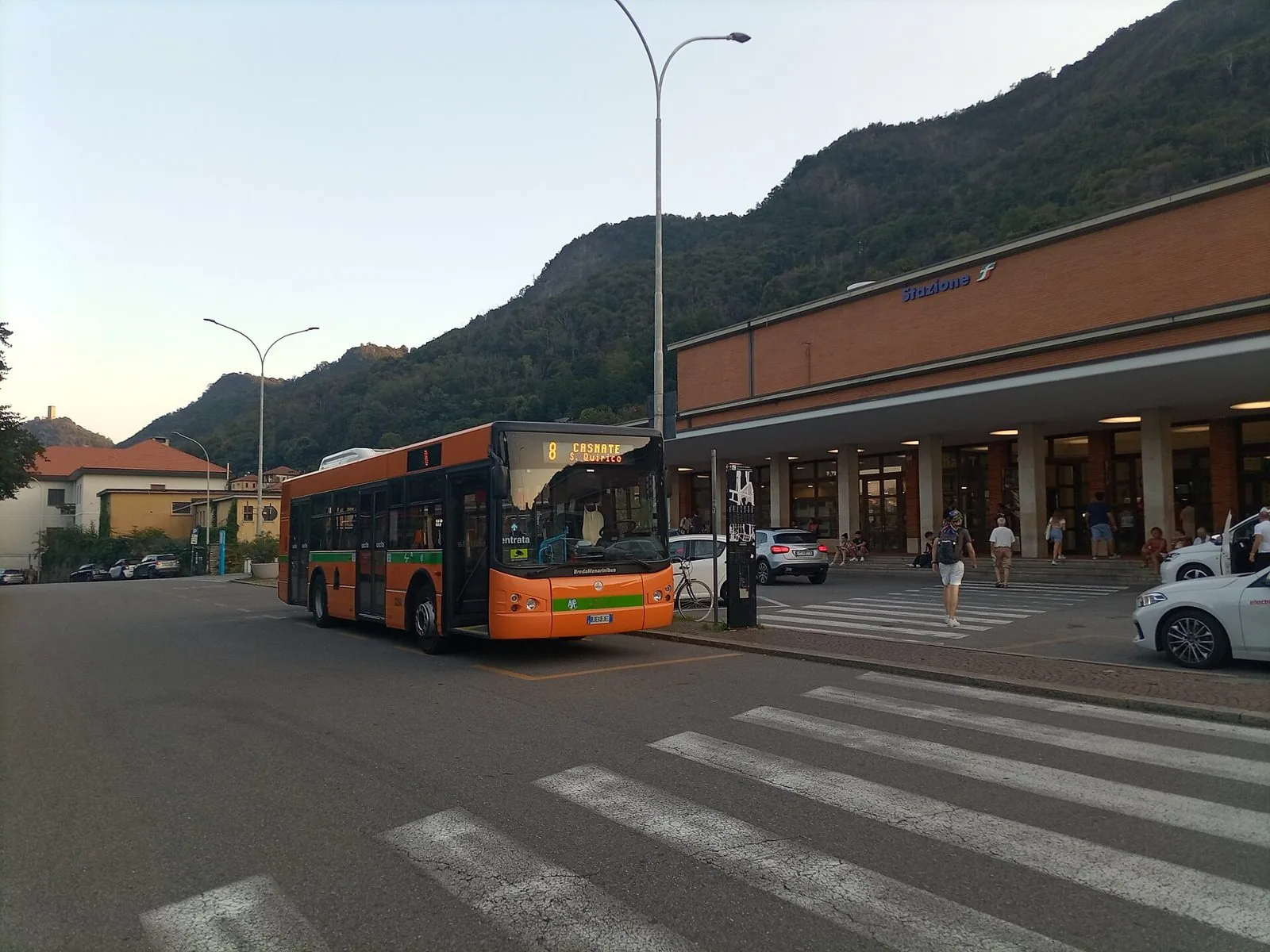 Il fabbricato della stazione ferroviaria di Como San Giovanni con un autobus urbano arancione fermo davanti e la collina boscosa sullo sfondo