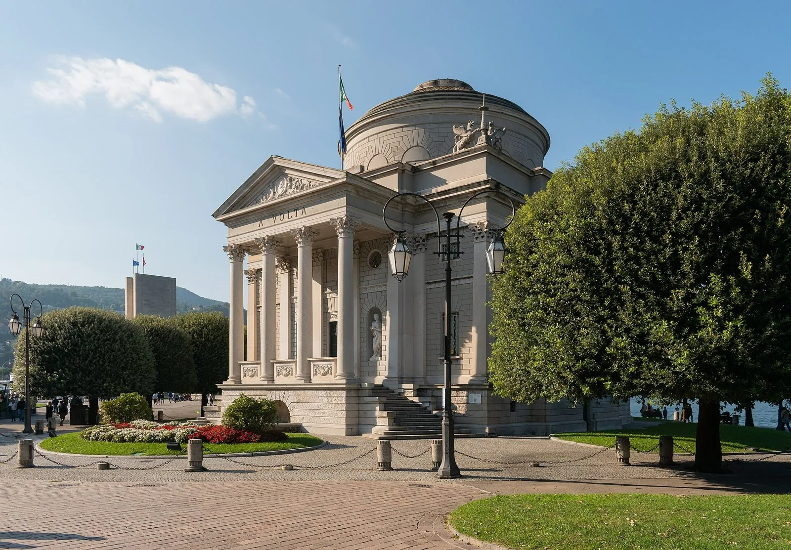 Il Tempio Voltiano di Como, edificio neoclassico con colonne e cupola, circondato da aiuole fiorite e alberi sotto un cielo azzurro