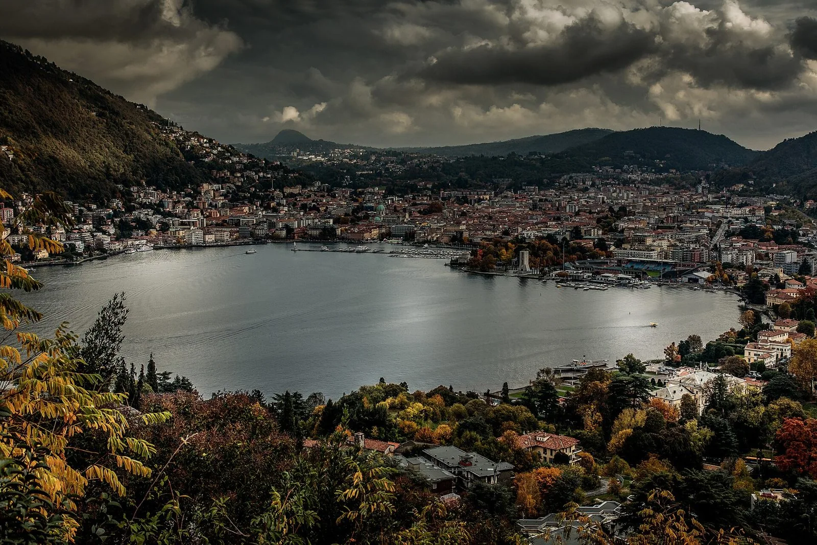 Panorama autunnale di Como e del primo bacino del lago visto dall'alto, con alberi dalle foglie gialle e arancioni in primo piano e cielo nuvoloso
