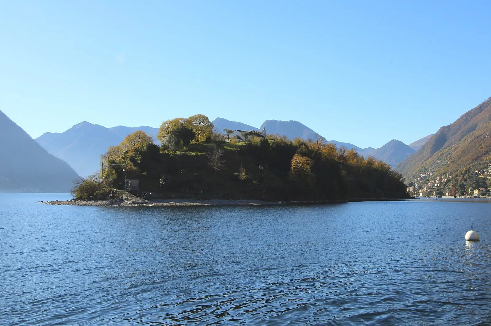 L'Isola Comacina vista dal lago, coperta di alberi autunnali con una piccola chiesa sulla sommità e le montagne sullo sfondo